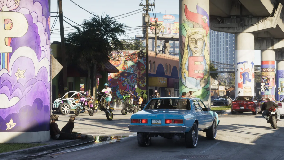 Captura oficial de GTA VI: un coche clásico azul circula bajo un paso elevado con columnas pintadas en Vice City, entre motoristas y vecinos reunidos junto a la calzada.