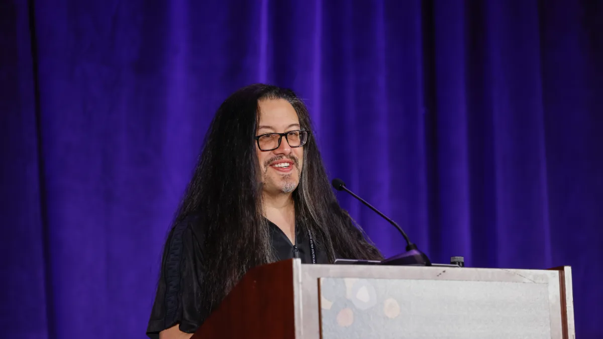 John Romero, con gafas y larga melena oscura, sonríe mientras habla en un atril con micrófono ante una cortina violeta durante la Game Developers Conference de 2022.
